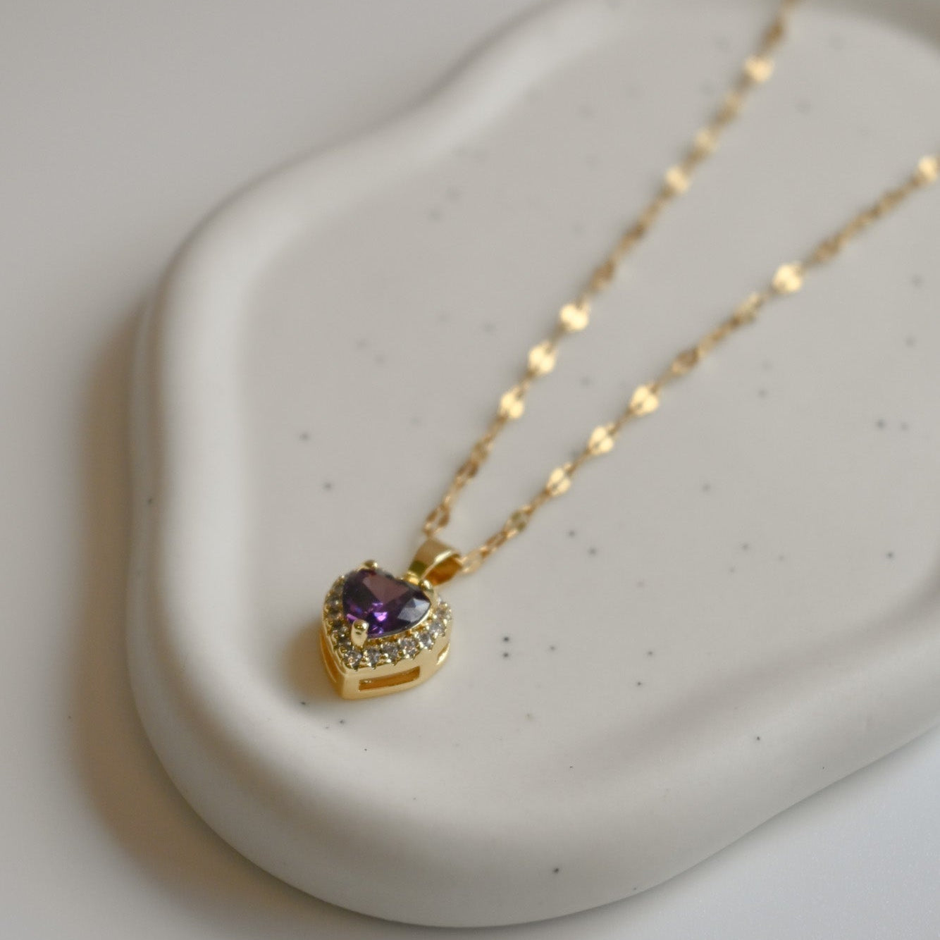 Gold necklace with a heart-shaped pendant featuring an amethyst stone on a white background