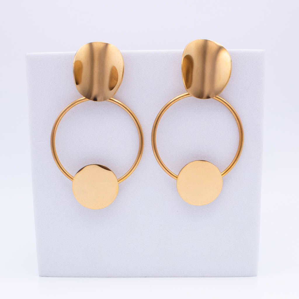 A pair of gold-toned statement earrings with a flat oval stud, a thin circular hoop, and a flat circular disc dangling below, displayed on a white rectangular stand against a white background.