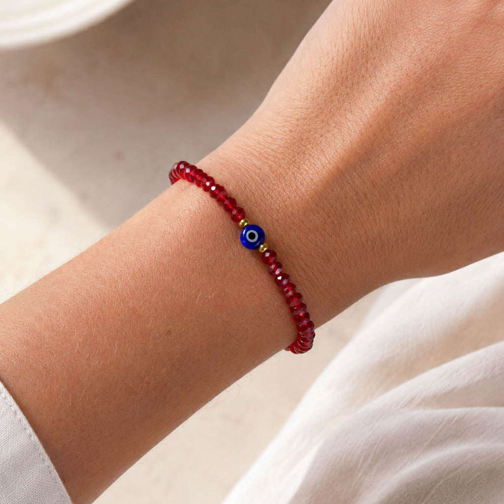 A close-up of a person's wrist wearing a red beaded bracelet with a blue and white evil eye charm and small gold spacer beads.