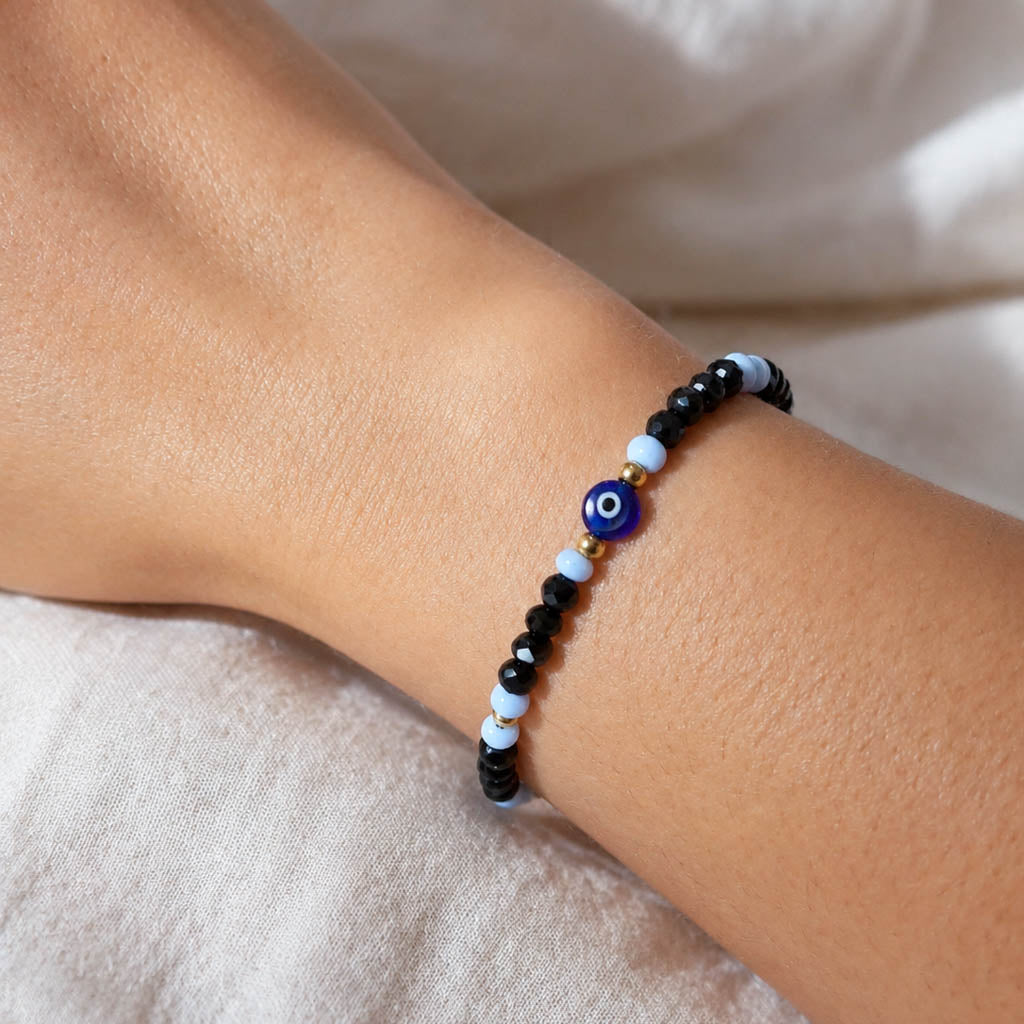 A close-up of a beaded bracelet worn on a person's wrist. The bracelet features black beads, light blue beads, and a central blue and white evil eye charm with a small gold accent.
