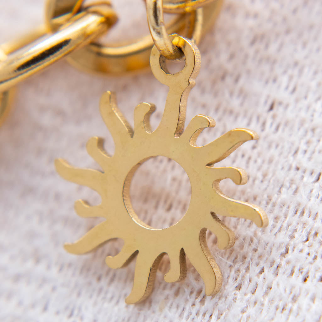 A close-up of a gold-toned sunburst charm with a cutout center, attached to a matching chain, resting on a textured off-white fabric background.