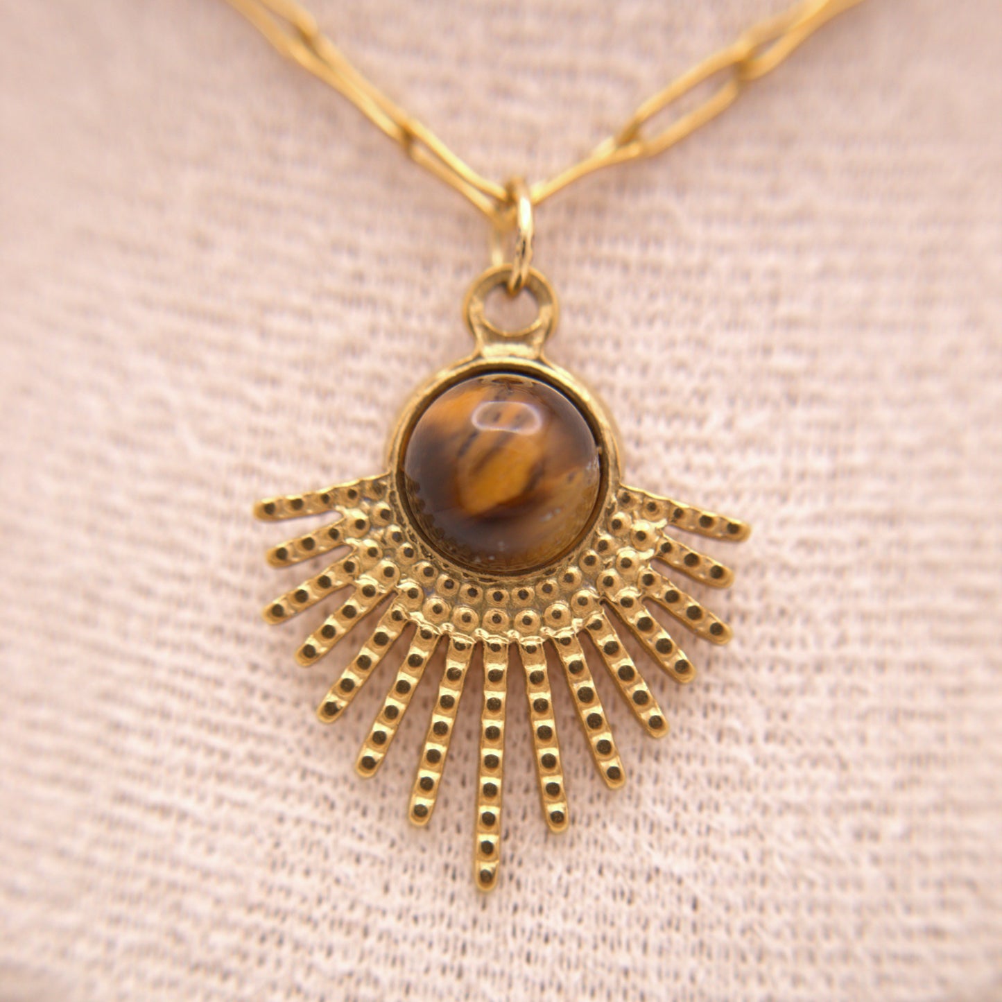 Gold necklace with a sunburst pendant featuring a tiger's eye stone on a beige fabric background