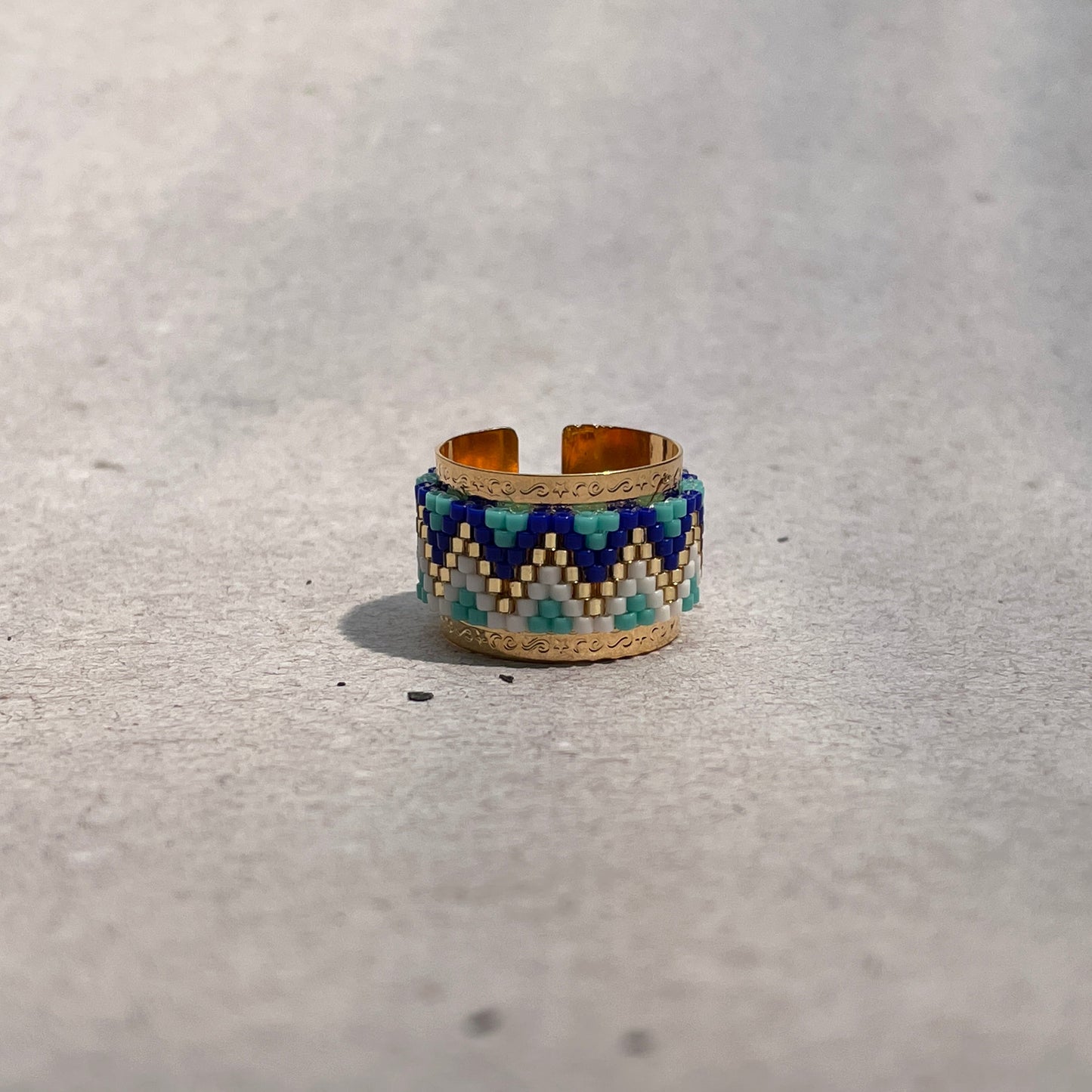 A handcrafted bronze ring with blue, gold, and red geometric patterns on the top surface, placed on a gray road surface.