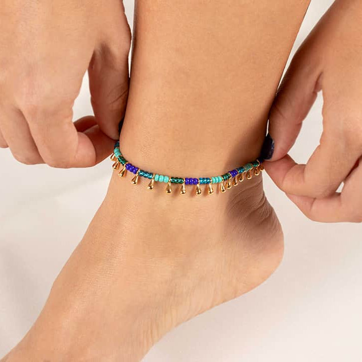 An adjustable ankle bracelet with blue and gold gold plated appliques, being worn on a person's ankle.