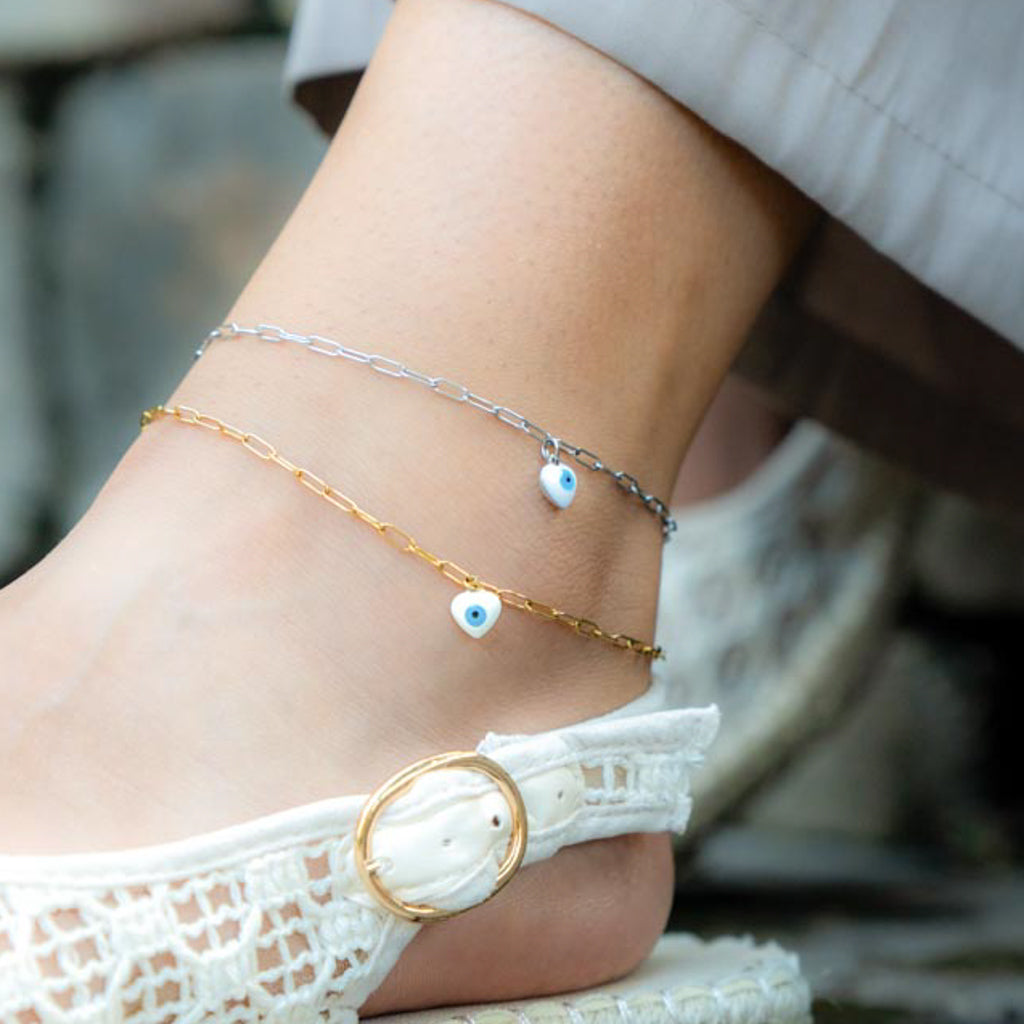 A close-up of a person's ankle wearing two delicate anklets: one gold chain with a small white heart charm containing a blue evil eye, and one silver chain with a similar heart charm. White lace flat sandals are also visible.