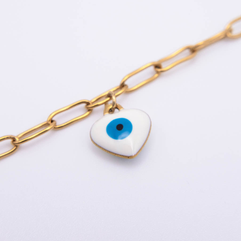 A gold paperclip chain anklet featuring a small white enamel evil eye charm with a blue iris and black pupil, set against a plain white background.