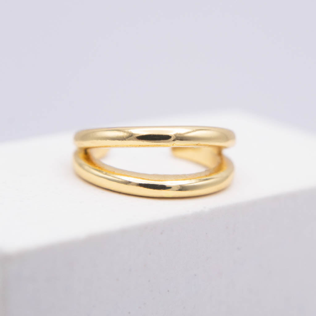 A polished gold ear cuff with a double-loop, intertwined design, resting on a white textured block against a light gray background.