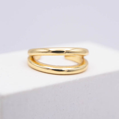 A polished gold ear cuff with a double-loop, intertwined design, resting on a white textured block against a light gray background.