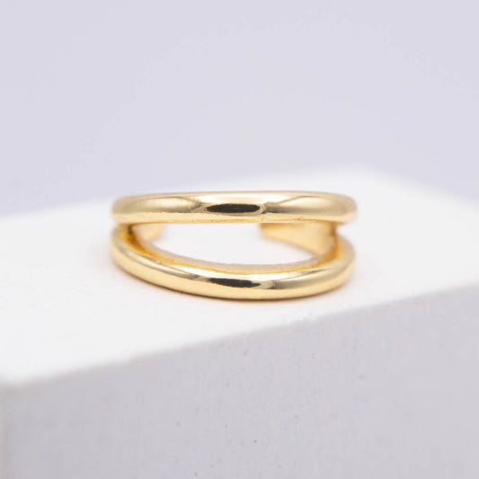 A polished gold ear cuff with a double-loop, intertwined design, resting on a white textured block against a light gray background.