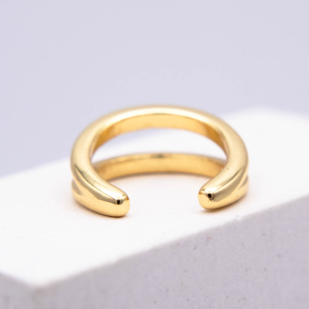 A polished gold wrap ear cuff displayed on a white textured block against a soft light gray background.