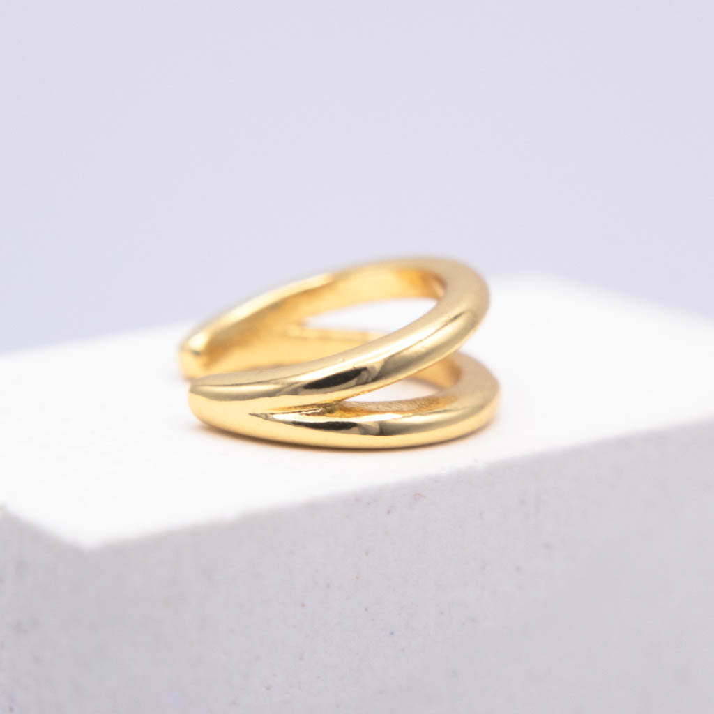 A polished gold wrap ear cuff displayed on a white textured block against a soft light gray background.
