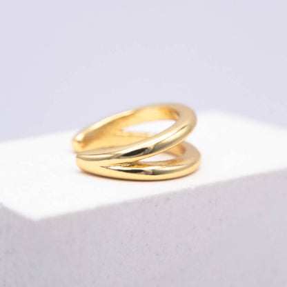 A polished gold wrap ear cuff displayed on a white textured block against a soft light gray background.