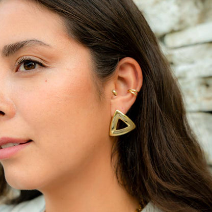 A close-up of a woman's ear wearing a gold triangular hoop earring, with matching small gold ear cuffs, against a softly blurred light stone background.