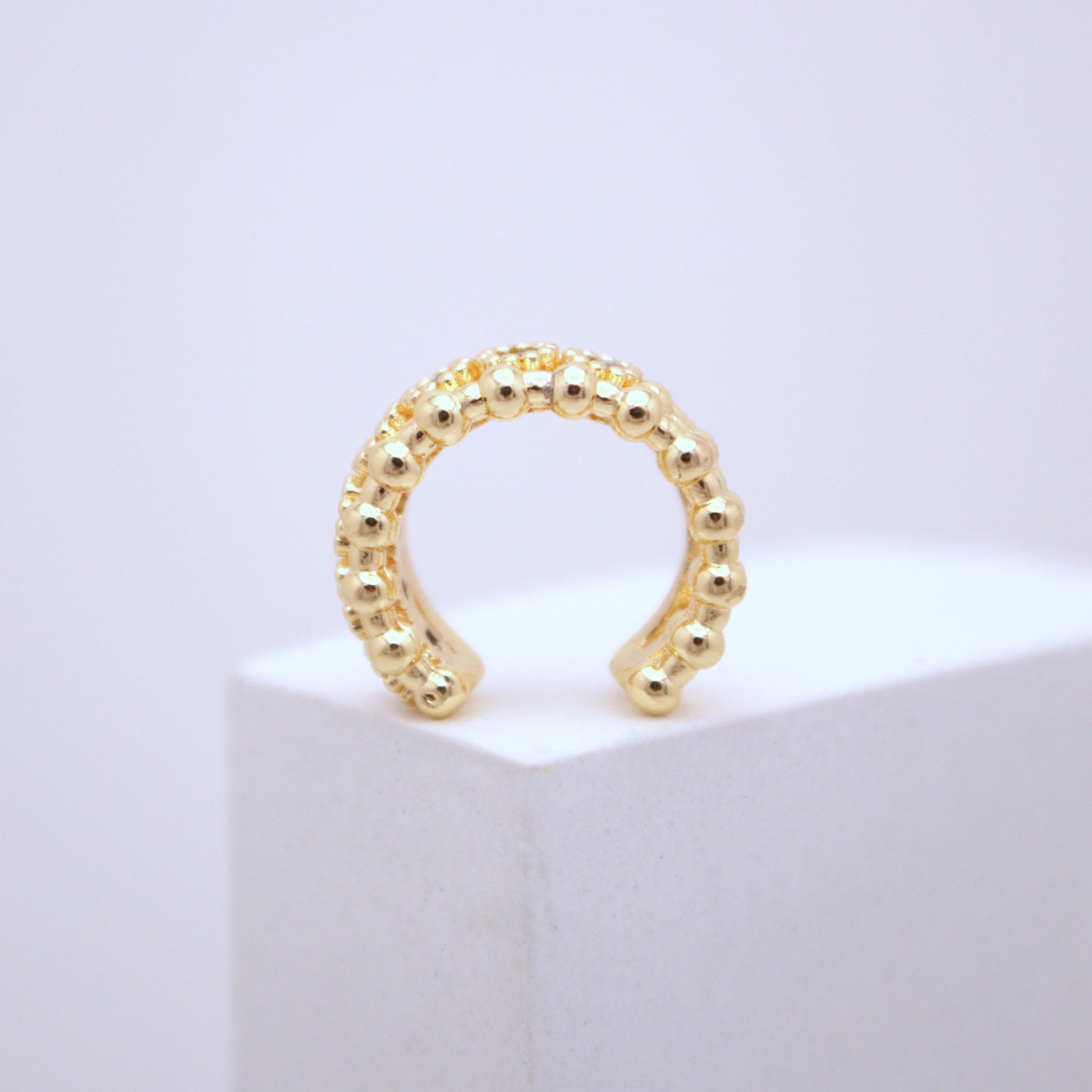 Gold beaded ear cuff on a white pedestal with a light purple background