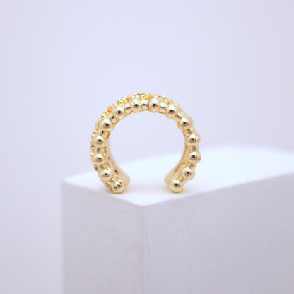 Gold beaded ear cuff on a white pedestal with a light purple background