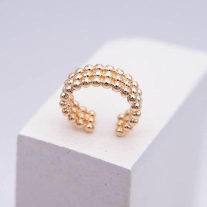 A gold beaded open ring with a textured, spherical bead pattern, displayed on a white geometric block against a light gray background.
