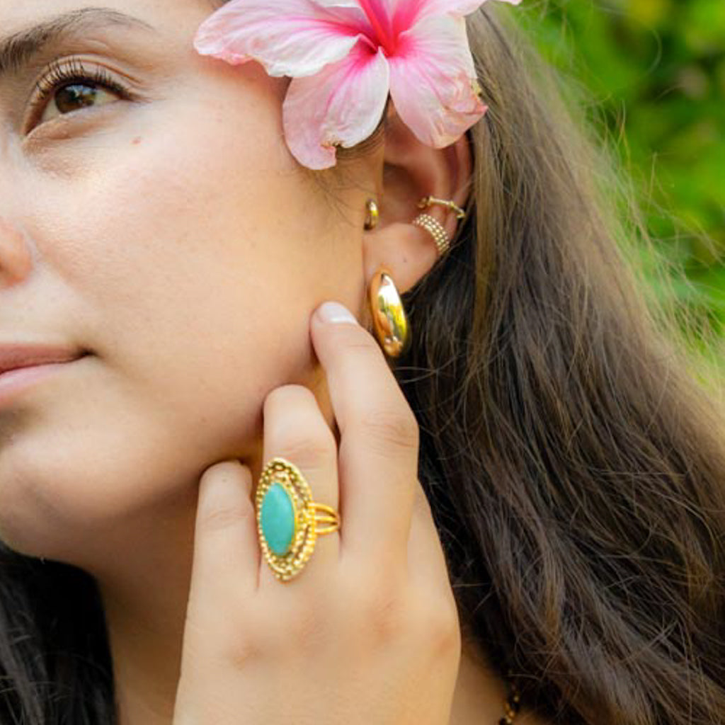 A close-up lifestyle shot of a woman wearing gold jewelry, featuring a gold ring with a turquoise oval stone, a gold hoop earring, and a gold ear cuff, with a pink hibiscus flower in her hair against a green outdoor background.