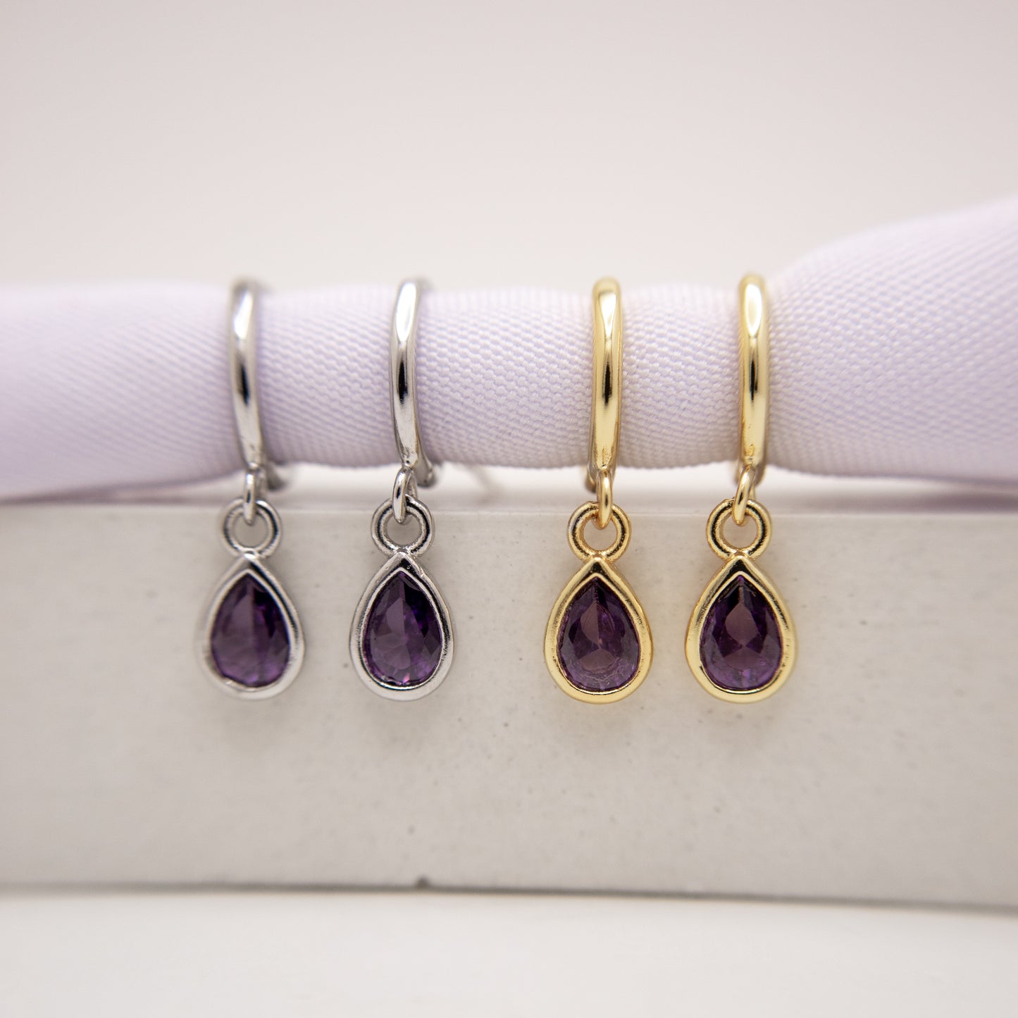 Two pairs of teardrop-shaped amethyst earrings, one in silver and one in gold, on a white surface.