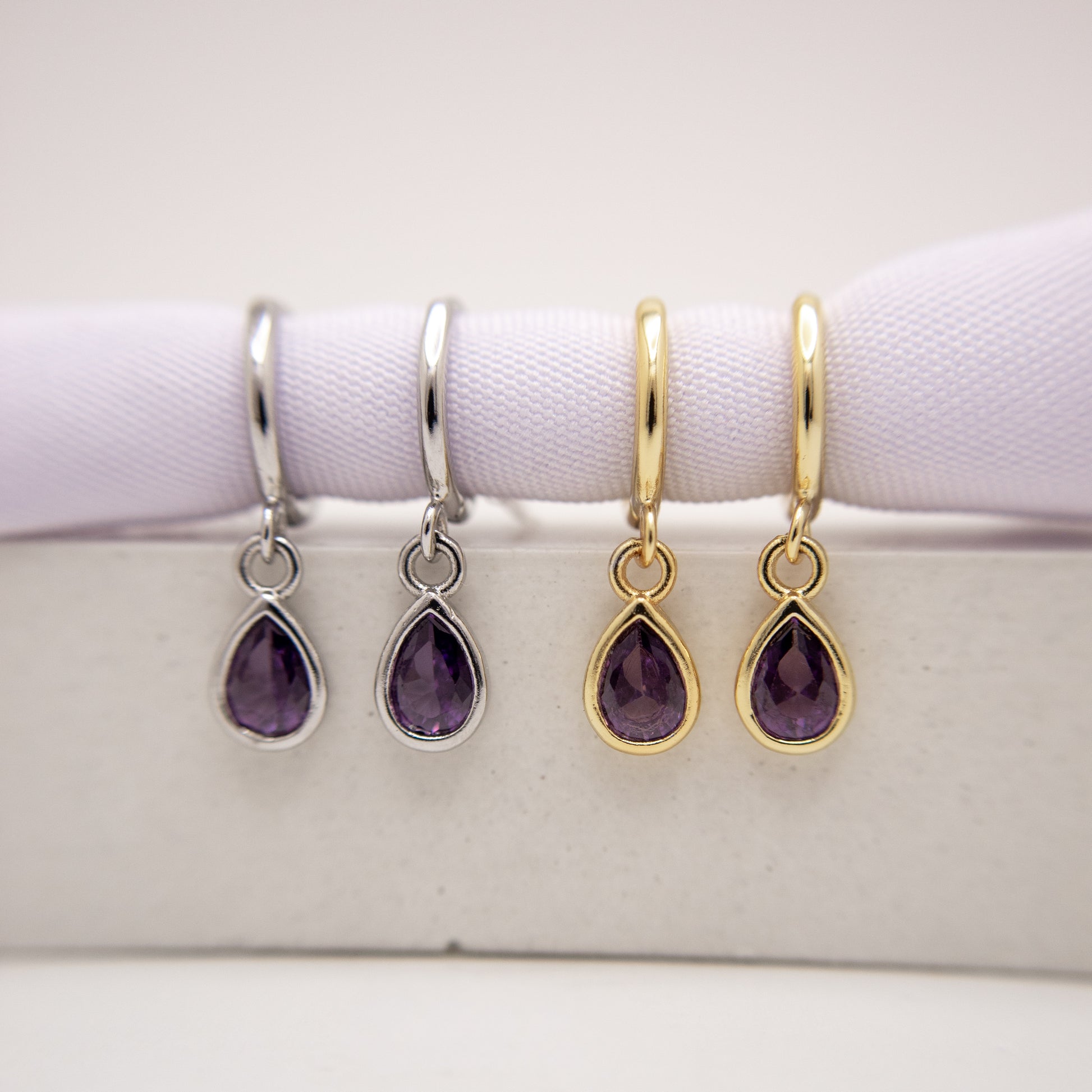 Two pairs of teardrop-shaped amethyst earrings, one in silver and one in gold, on a white surface.