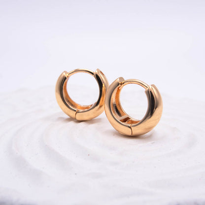 A pair of polished gold huggie hoop earrings resting on a textured white surface against a soft white background.
