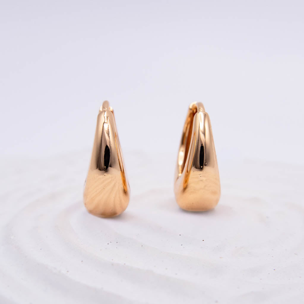 A pair of polished gold hoop earrings with a teardrop-shaped, textured surface, displayed on a white textured surface against a light gray background.