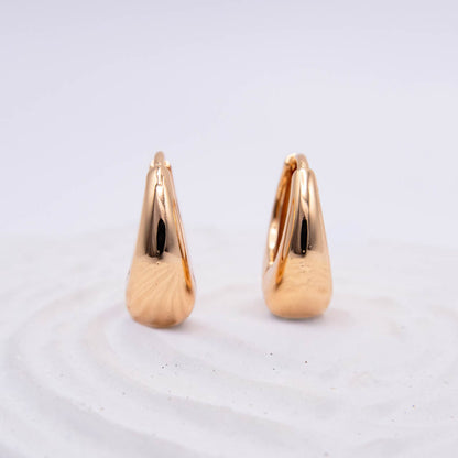 A pair of polished gold hoop earrings with a teardrop-shaped, textured surface, displayed on a white textured surface against a light gray background.