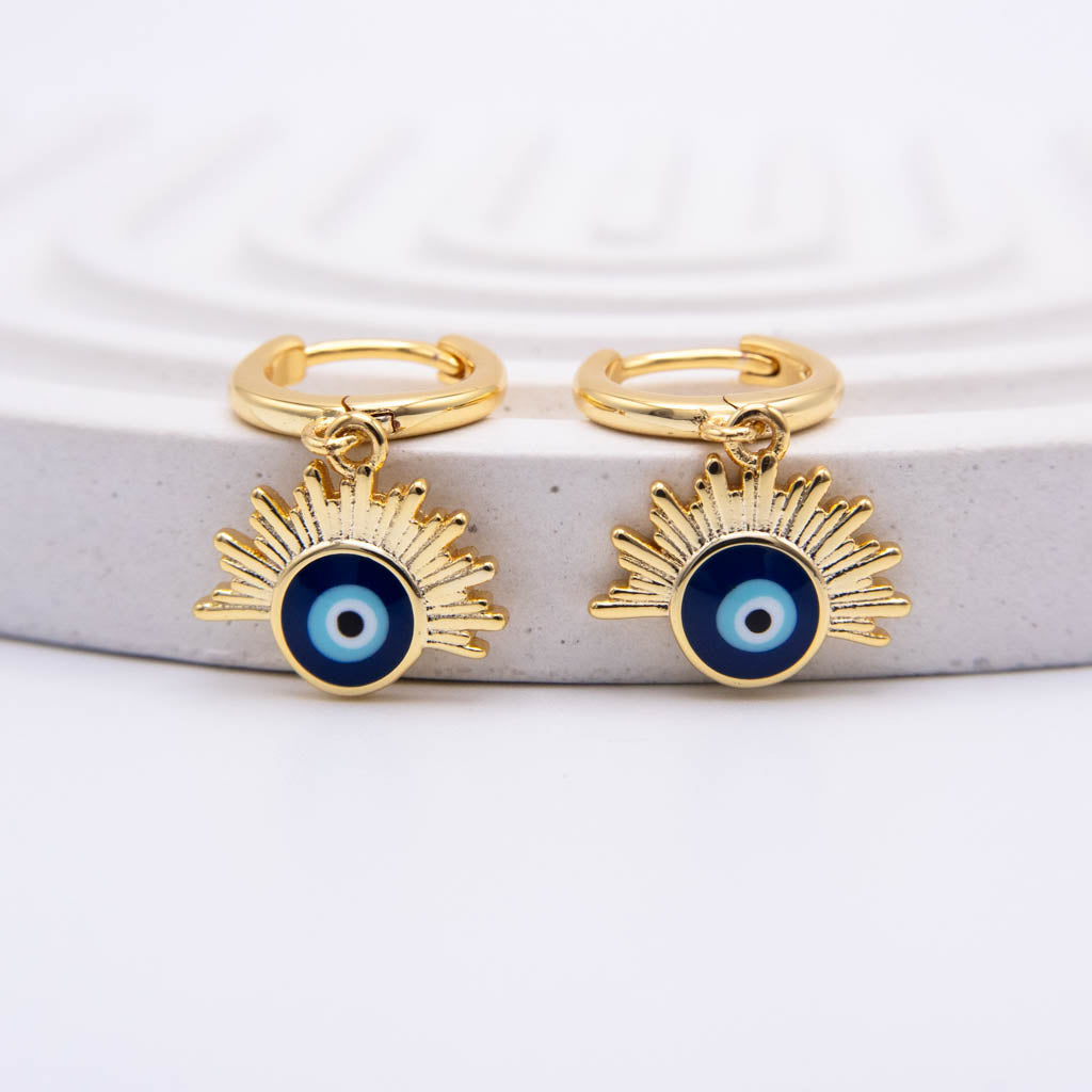 A pair of gold huggie hoop earrings with dangling sunburst charms featuring a blue and white evil eye design, displayed on a light gray textured stone riser against a white background.