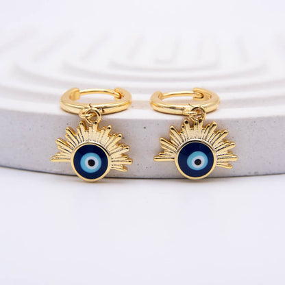 A pair of gold huggie hoop earrings with dangling sunburst charms featuring a blue and white evil eye design, displayed on a light gray textured stone riser against a white background.