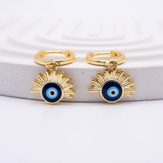 A pair of gold huggie hoop earrings with dangling sunburst charms featuring a blue and white evil eye design, displayed on a light gray textured stone riser against a white background.