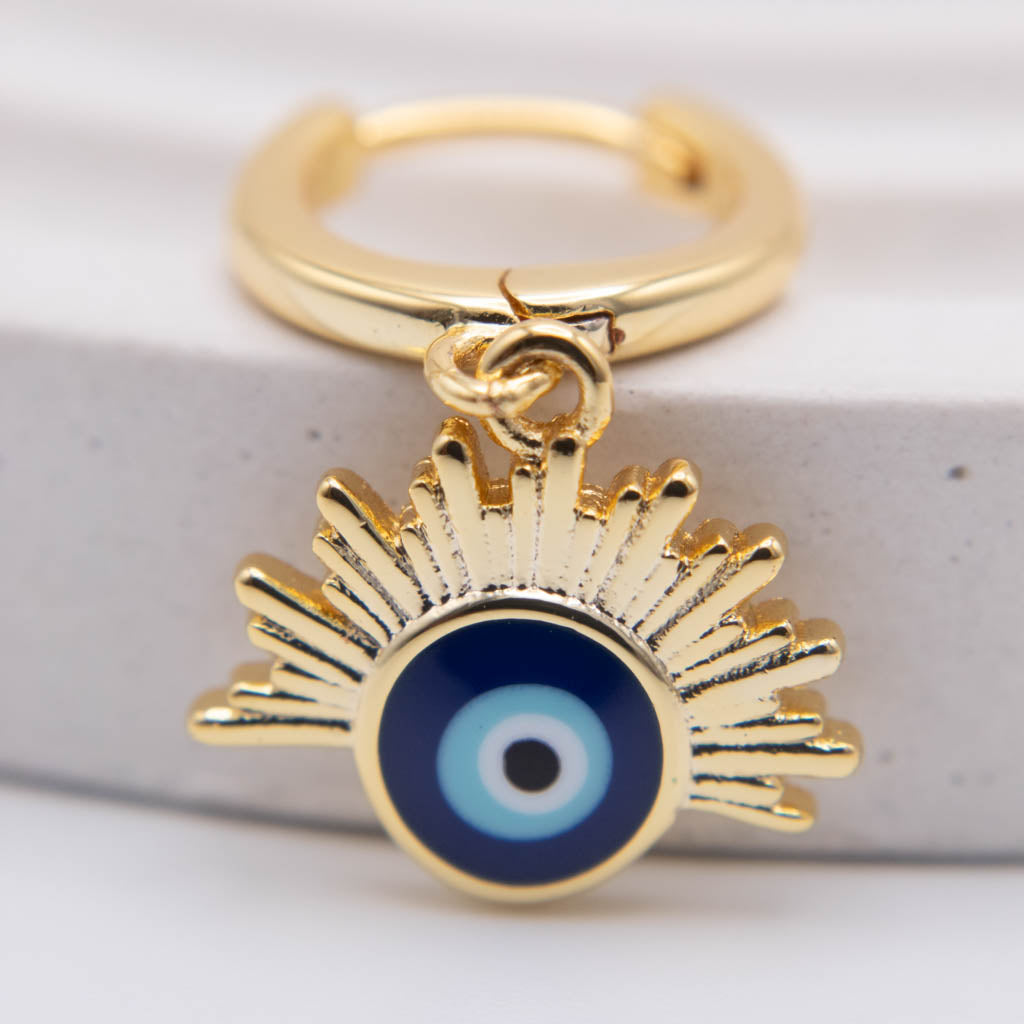 Gold huggie hoop earring with a dangling evil eye charm featuring blue, white, and black enamel details, set against a light gray textured background.