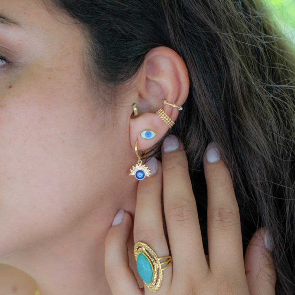 A close-up of a person's ear and hand showcasing multiple gold ear piercings and rings. The ear features a small hoop, a gold ear cuff with an evil eye motif, a gold spiral ear clip, and a gold dangle earring with a sunburst halo and blue evil eye center.