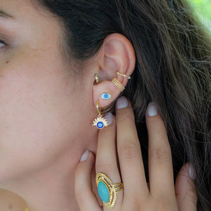 A close-up of a person's ear and hand showcasing multiple gold ear piercings and rings. The ear features a small hoop, a gold ear cuff with an evil eye motif, a gold spiral ear clip, and a gold dangle earring with a sunburst halo and blue evil eye center.
