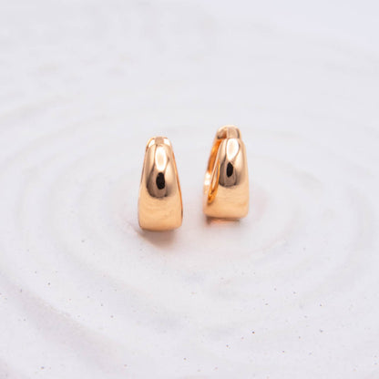 A pair of small polished rose gold hoop earrings standing upright on a light textured white surface.