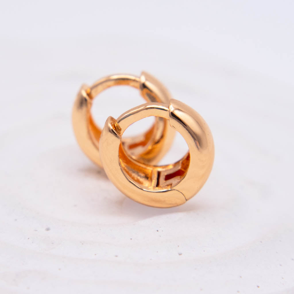 A pair of polished gold huggie hoop earrings on a light, off-white textured background.