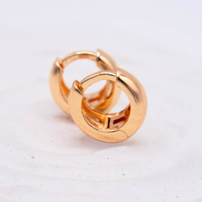 A pair of polished gold huggie hoop earrings on a light, off-white textured background.
