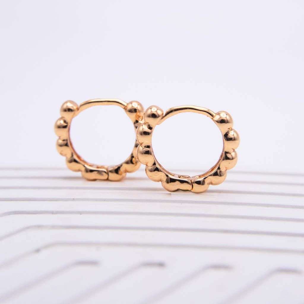 A pair of gold-toned hoop earrings with a scalloped, beaded border, displayed on a white ribbed surface against a white background.