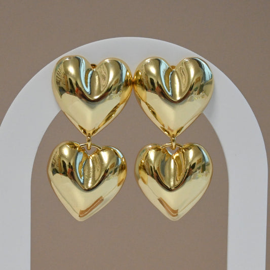 A pair of gold-colored double heart-shaped stud earrings hanging from a white metal stud base.