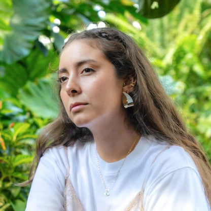 A woman with long brown hair wearing a white t-shirt, a delicate gold necklace, and a silver geometric hoop earring, set against a lush green background of tropical foliage.