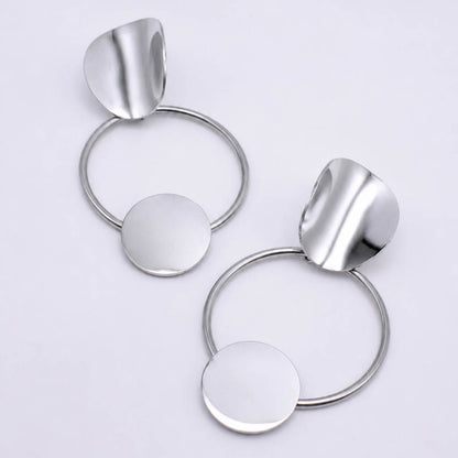 A pair of modern silver-toned statement earrings featuring a circular ring, a flat round disc, and a curved leaf-like top piece, displayed on a clean white background.