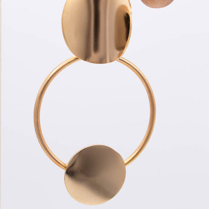 A close-up of a gold-toned geometric earring featuring a large circular hoop with a flat disc pendant and a polished oval stud, shown against a clean white background.