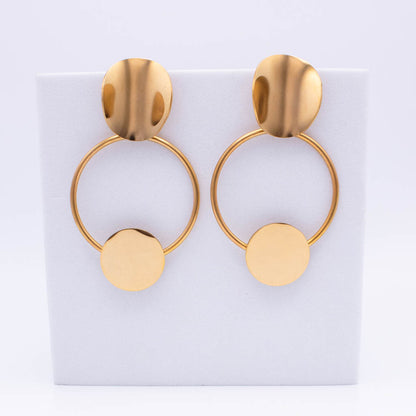 A pair of gold-toned statement earrings with a flat oval stud, a thin circular hoop, and a flat circular disc dangling below, displayed on a white rectangular stand against a white background.