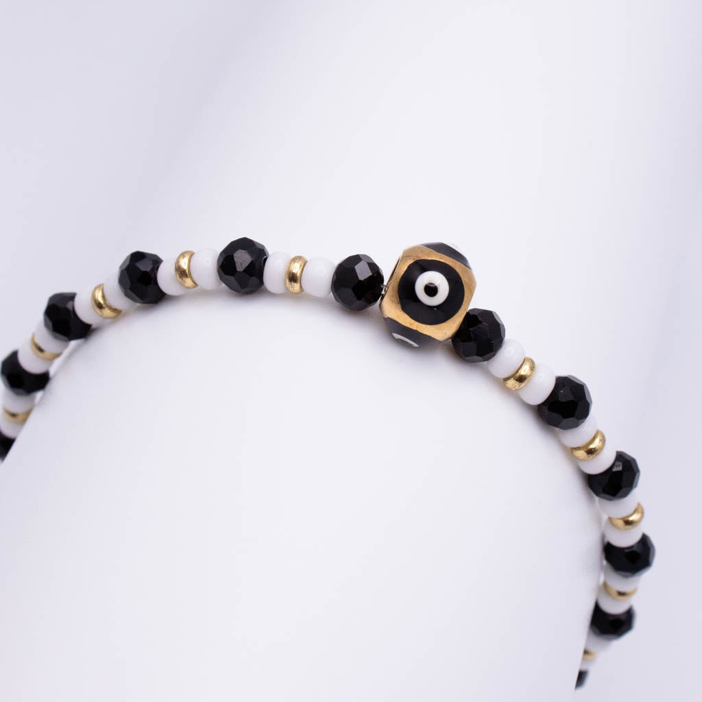 A beaded bracelet with alternating black faceted beads and white beads, accented by small gold spacer beads and a central gold evil eye charm, displayed on a white mannequin arm.
