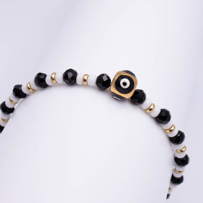 A beaded bracelet with alternating black faceted beads and white beads, accented by small gold spacer beads and a central gold evil eye charm, displayed on a white mannequin arm.