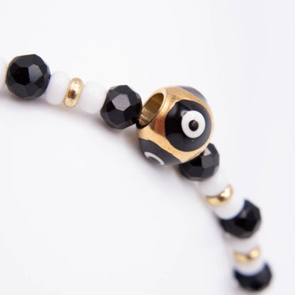 A close-up of a beaded bracelet featuring black faceted beads, white beads, small gold spacer beads, and a central gold-toned evil eye charm with black, white, and red detailing, set against a plain white background.