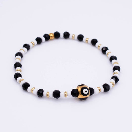 A beaded stretch bracelet featuring black faceted beads, white round beads, small gold spacer beads, and a central black and white evil eye charm with a gold border, shown on a plain white background.