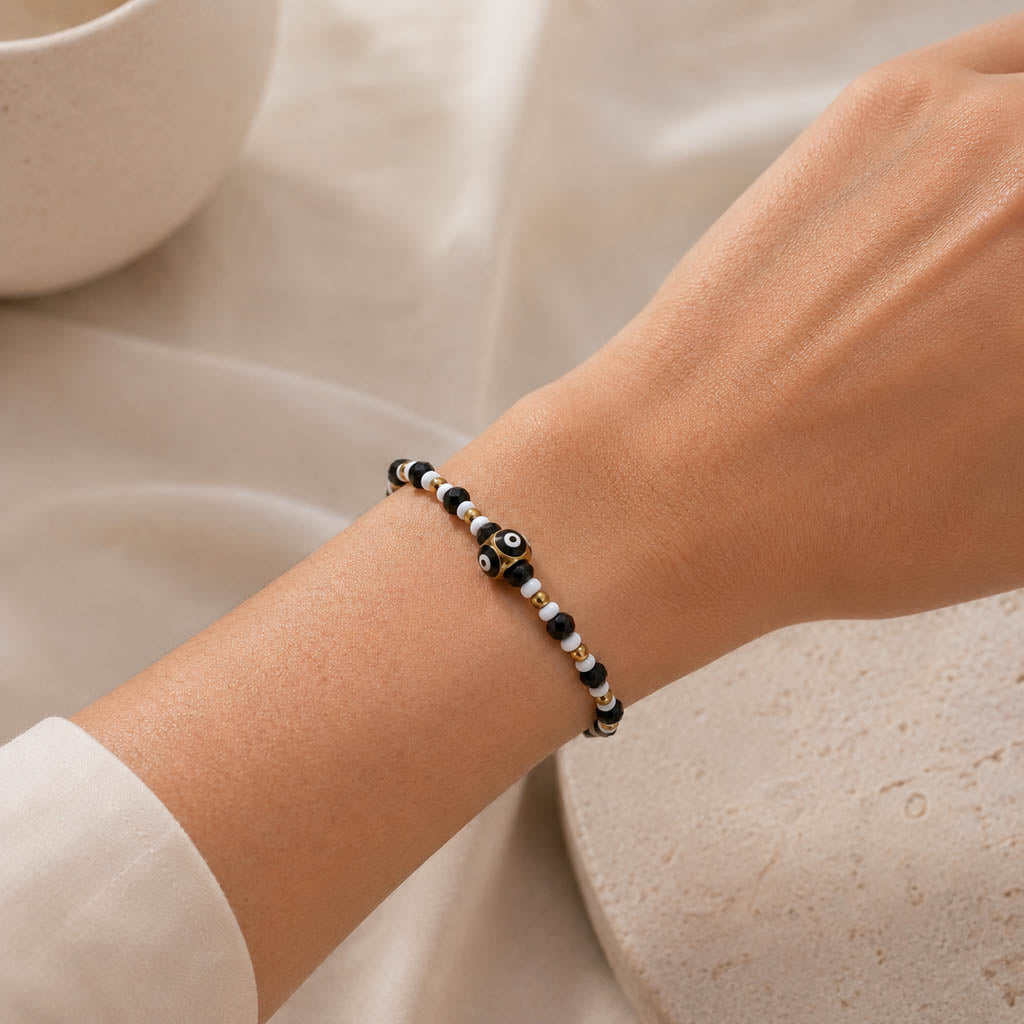 A close-up of a person's wrist wearing a beaded bracelet with black, white, and metallic gold beads, including a central evil eye charm.