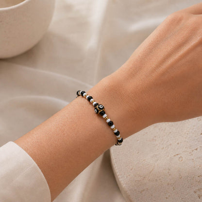 A close-up of a person's wrist wearing a beaded bracelet with black, white, and metallic gold beads, including a central evil eye charm.