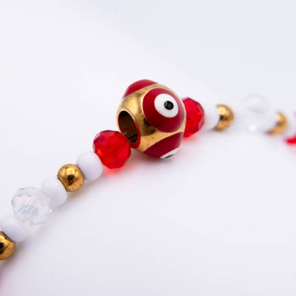 A close-up of a beaded bracelet featuring a central red and white evil eye bead with gold trim, flanked by red faceted beads, white round beads, and small gold metallic beads on a white background.
