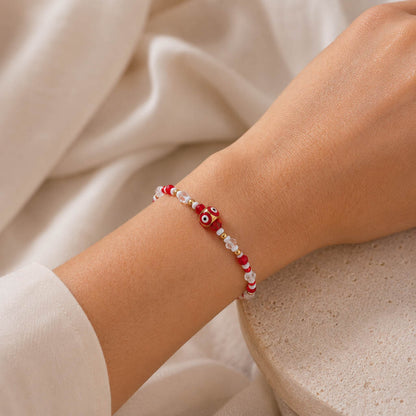 A close-up of a person's wrist wearing a delicate beaded bracelet with red and white beads and a central red evil eye charm, set against a soft cream fabric background.