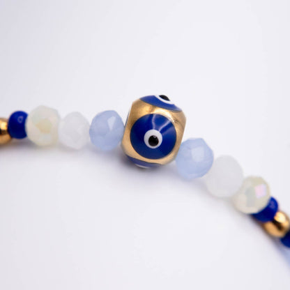 A close-up of a beaded strand featuring a central gold cube charm with a blue evil eye design, surrounded by faceted light blue, white, and dark blue beads on a white background.